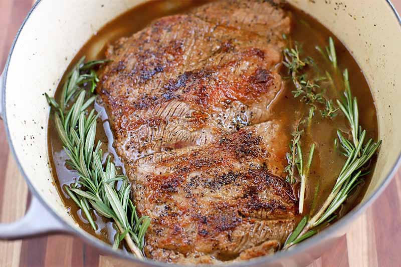 Brisket and fresh rosemary in a large enameled cooking pot, on a brown wood background.