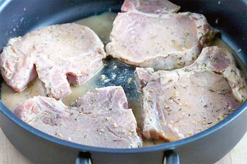 Four bone-in pork chops cooking in a large frying pan, in a cider marinade mixture.