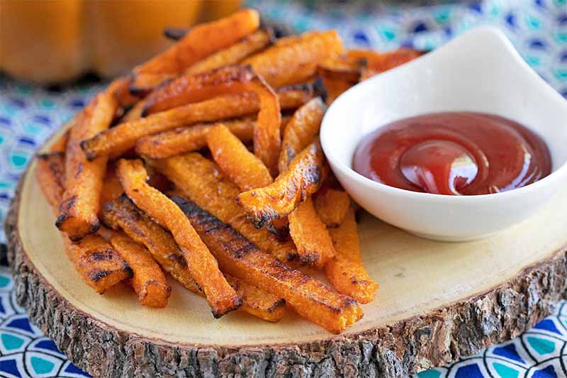 Baked butternut squash fries piled on a white plate with a small white bowl of ketchup.