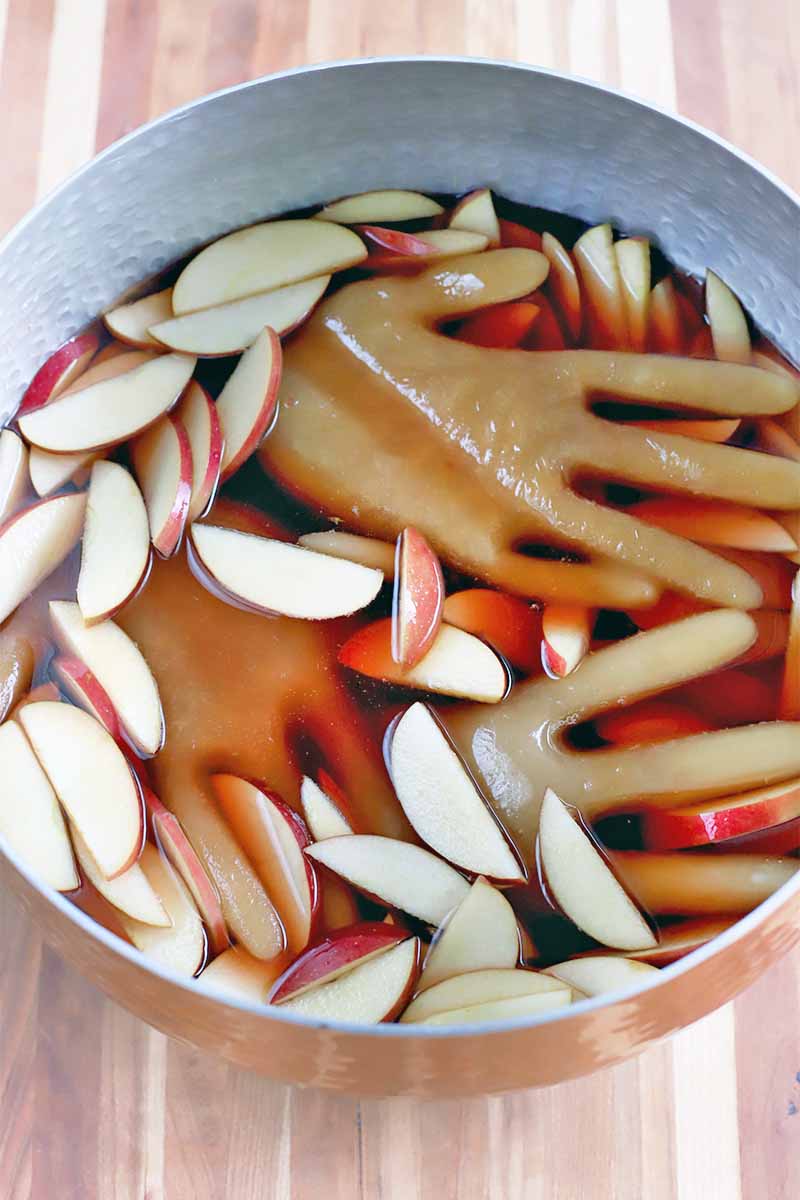 Top-down shot of a metal bowl of punch, with floating sliced fruit and peach-colored hand-shaped ice cubes, on a striped wood surface.