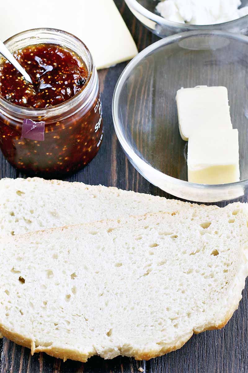 Top-down shot of two slices of sourdough bread, a jar of jam with a spoon stuck into it, a block of gruyere, a small bowl of goat cheese, and another small bowl of two chunks of butter, on a brown wood background.