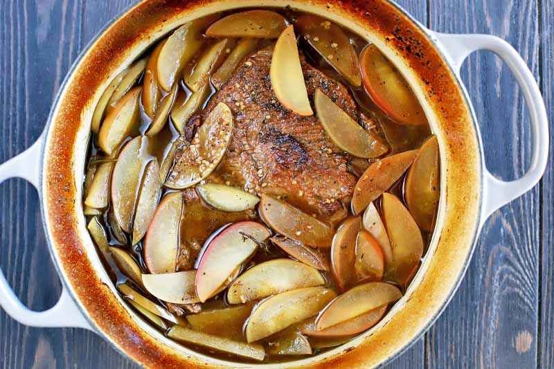 Top-down shot of a pot of brisket and sliced apples, on a dark brown wood background.