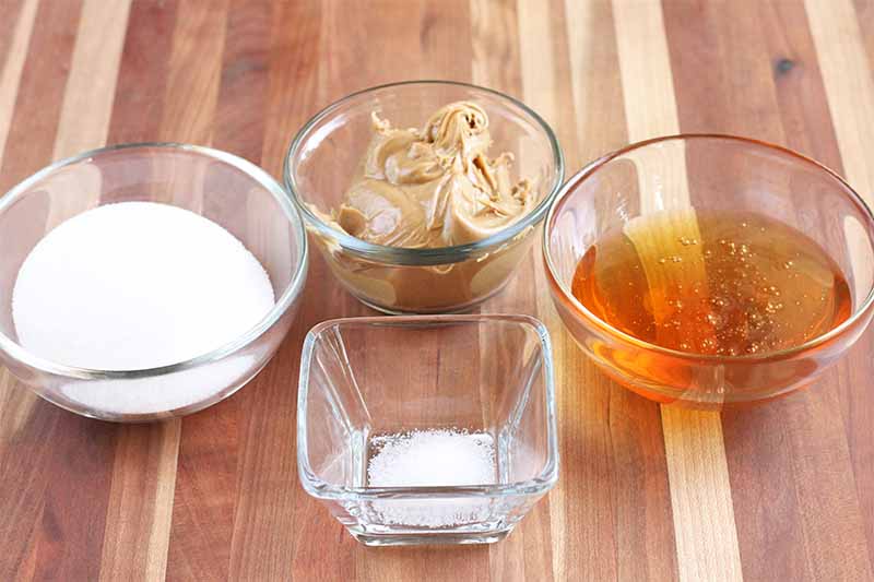 Three round glass dishes of sugar, peanut butter, and honey, in a row behind a small square glass dish of salt, on a striped brown wood surface.