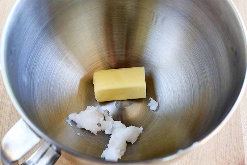 Half a stick of butter and a blob of solid coconut oil are at the bottom of a stainless steel stand mixer bowl, on a beige surface.