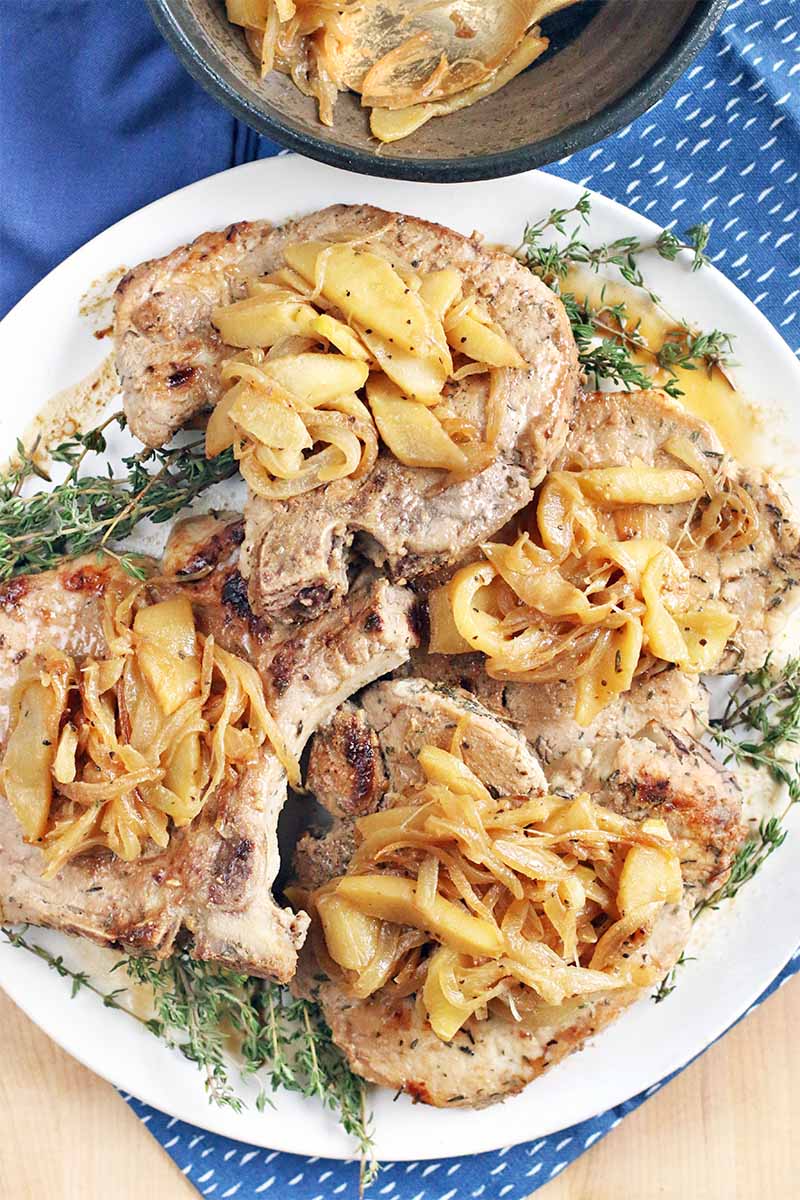 Top-down view of cider marinated pork chops topped with caramelized apples and onions, with sprigs of thyme for garnish, on a large plate on top of a blue cloth on a beige surface, with a gray bowl of more of the topping closely cropped at the top of the frame.