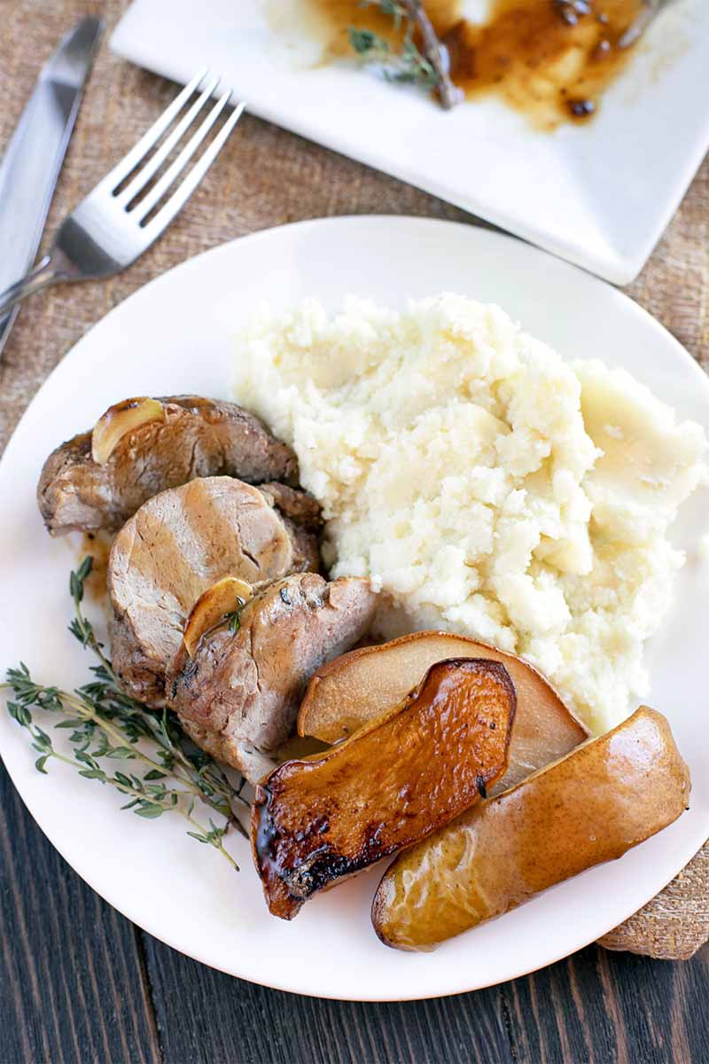Sliced pork tenderloin on a white dinner plate with sauteed pears, fresh thyme, and mashed potatoes, with silverware to the left, and a square serving platter at the top of the frame.