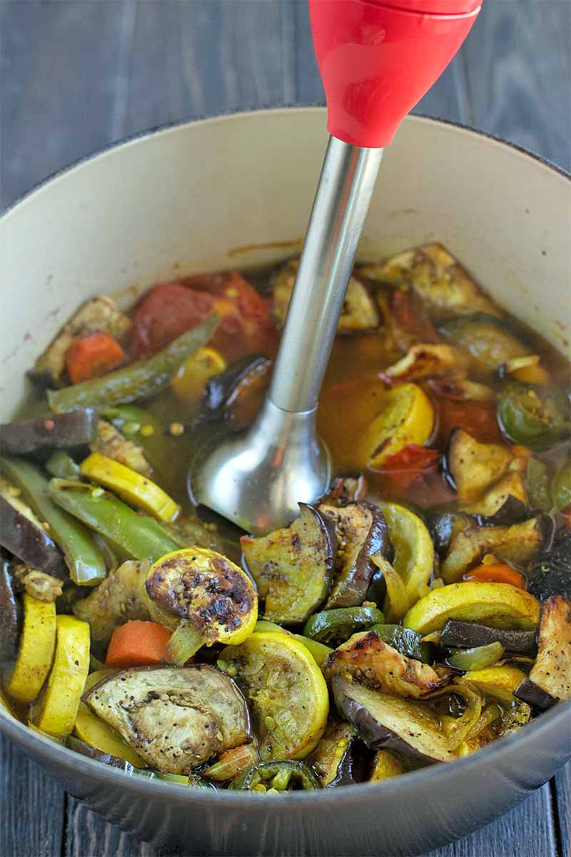 An immersion blender in an enameled stock pot full of cooked vegetables and brown broth.