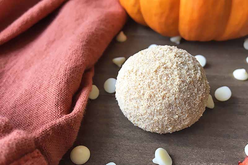 Horizontal image of a single truffle on a wooden surface by an orange towel, white chocolate chips, and a pumpkin.
