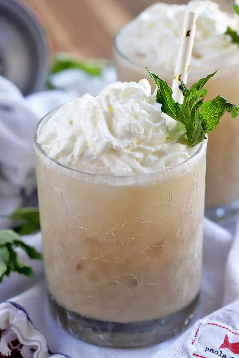 Closeup of a glass filled with a coffee-based cocktail and ice, topped with a swirl of whipped cream and a sprig of mint, with a white paper straw decorated with gold foil straws, with another identical beverage in the background, on a gathered white cloth dish towel with scattered fresh herbs, on a wood background.