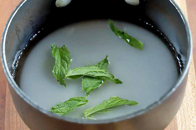 A nonstick saucepan of sugar, water, and mint leaves, on a brown wood surface.