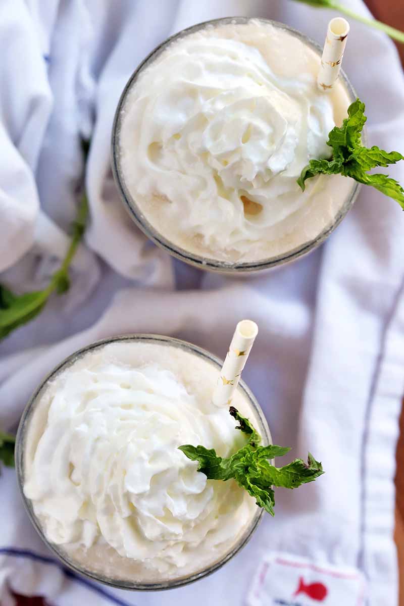 Overhead vertical shot of two glasses topped with whipped cream and sprigs of mint, with white paper straws decorate with gold foil straws, o a folded and gathered white cloth, on a brown wood surface.