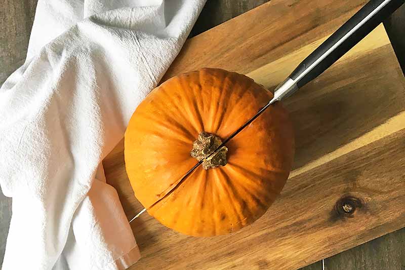 Horizontal image of a knife cutting through a small pumpkin on a wooden cutting board next to a white towel.