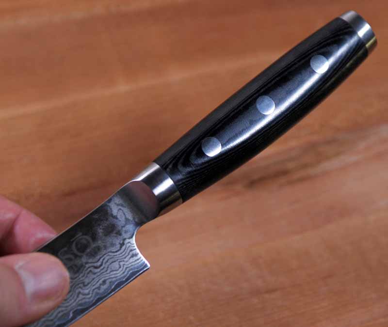 A human hand grasped the blade of the Enso HD Hammered Damascus Petty Utility Knife to show the black canvas micarta handle detail and polished end cap.