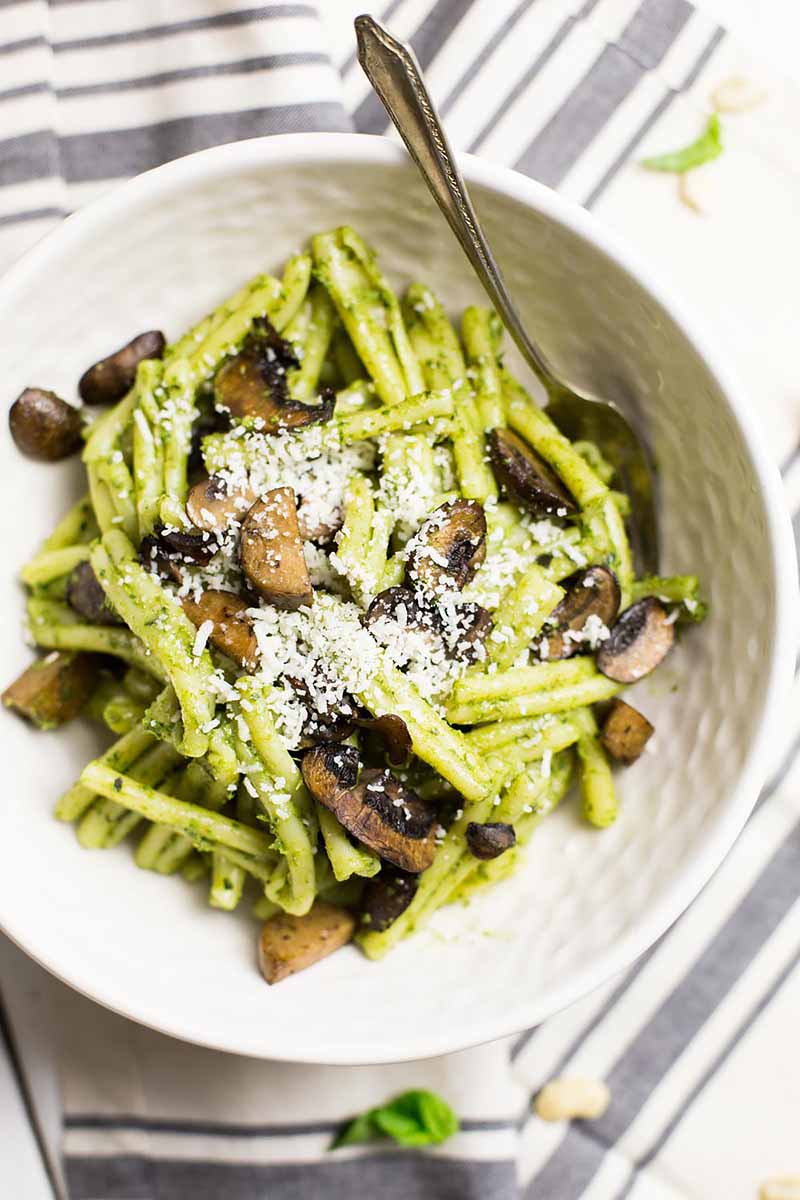 Vertical image of pasta mixed with mushrooms and a green sauce in a white bowl with a fork.