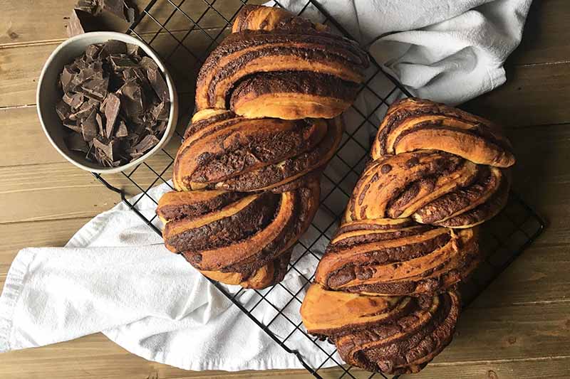 Horizontal image of two loaves of baked pastry on a cooling rack next to a bowl of chocolate.