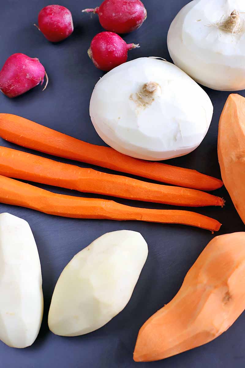 Peeled carrots, potatoes, turnips, sweet potatoes, and a few whole radishes, on a slate surface.