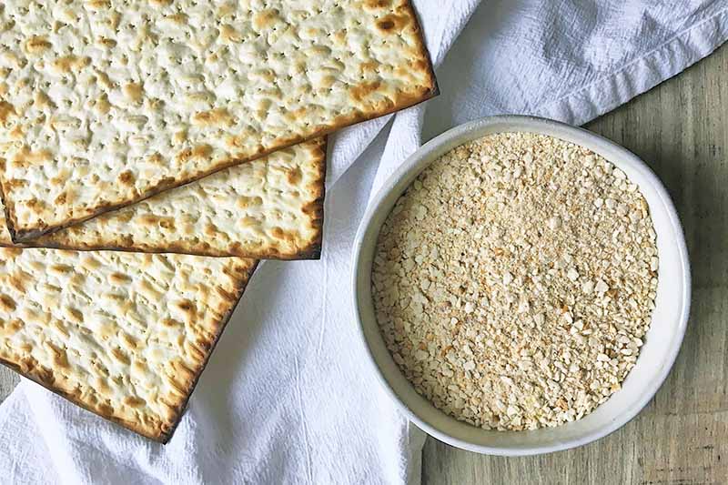 Horizontal image of matzo meal in a bowl next to the whole crackers on a white towel.