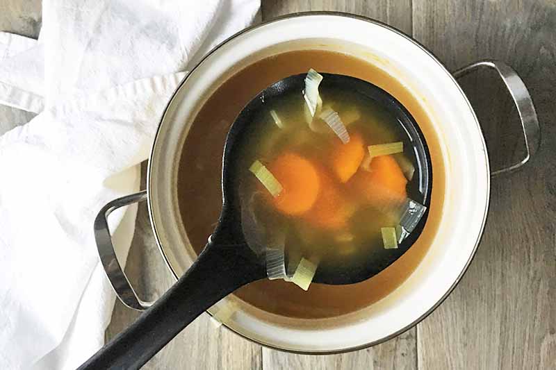 Horizontal image of a black spoon with carrots and leeks in a liquid over a pot.