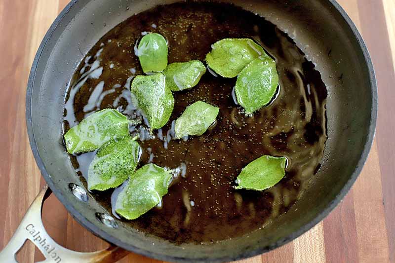 Sage leaves frying in oil in a saucepan, on a brown striped wood background.