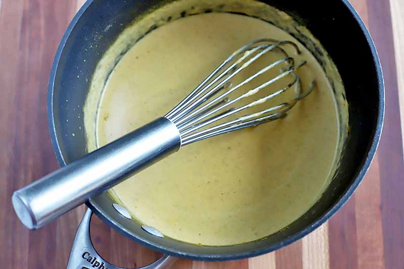 A nonstick saucepan of pumpkin cream sauce, with a wire whisk, on a striped beige and brown wood surface.