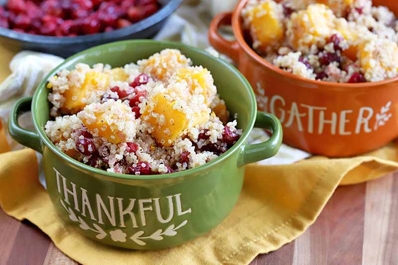 Two Thanksgiving-themed orange and green double-handled ceramic crocks of quinoa with winter squash and cranberries, on a yellow cloth on top of a wood surface, with a blue bowl of roasted berries on a beige cloth in the background.