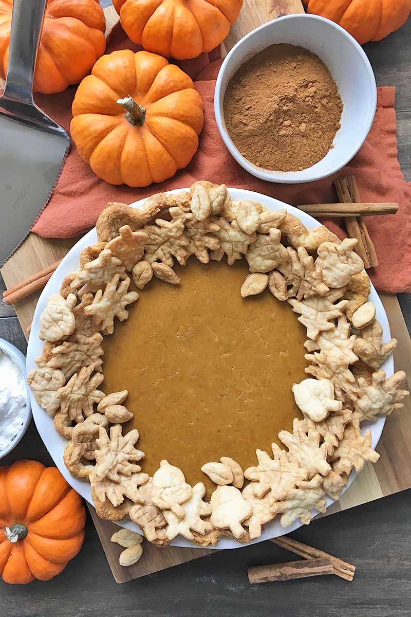 Vertical image of a whole pumpkin pie with a bowl of spices and decorations on an orange towel.