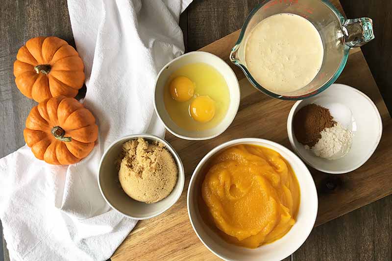 Horizontal image of ingredients in various bowls with pumpkins and a white towel on the side.
