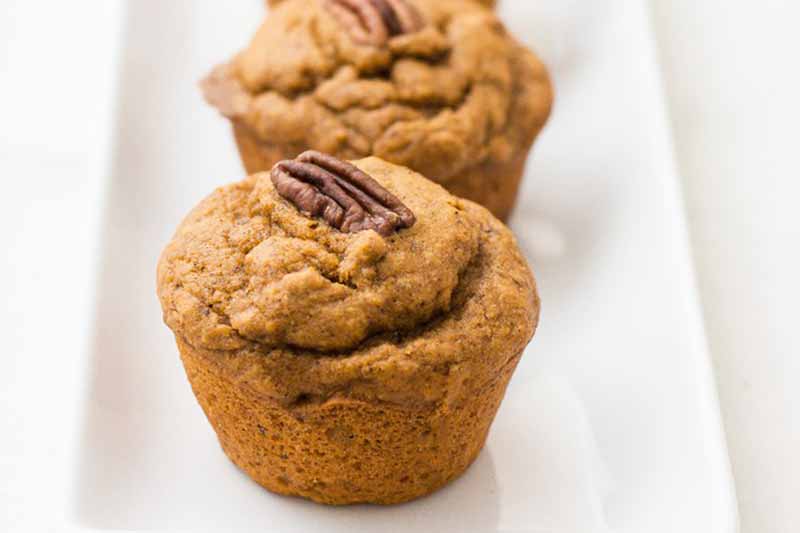 Horizontal close-up image of two pumpkin muffins with pecans on top of a white plate.