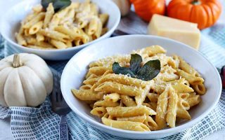 Two white bowls of pasta with pumpkin cream sauce, topped with grated cheese and fried sage leaves, on a blue and white checkered cloth with miniature white and orange pumpkins, and a block of Parmesan.