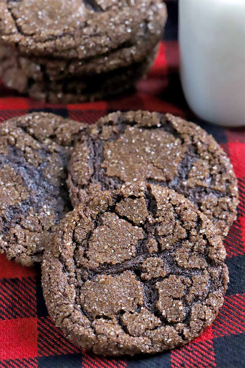 Three overlapping dark chocolate cookies fanned out on a surface topped with red and black plaid flannel fabric, with a short stack of the baked goods and a glass bottle of milk in the background.