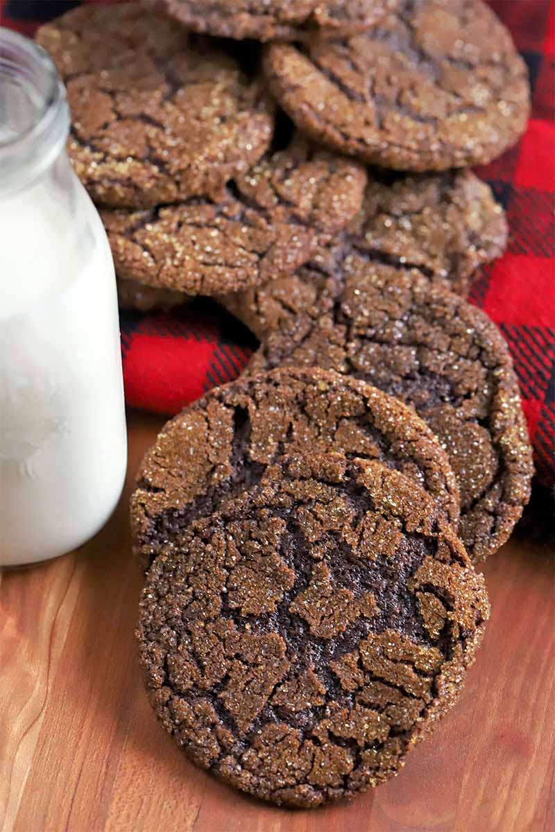 Homemade chocolate cookies are fanned out on a black and red piece of plaid flannel fabric and spilling out onto a brown wood surface, with a glass bottle of milk to the left of the frame.