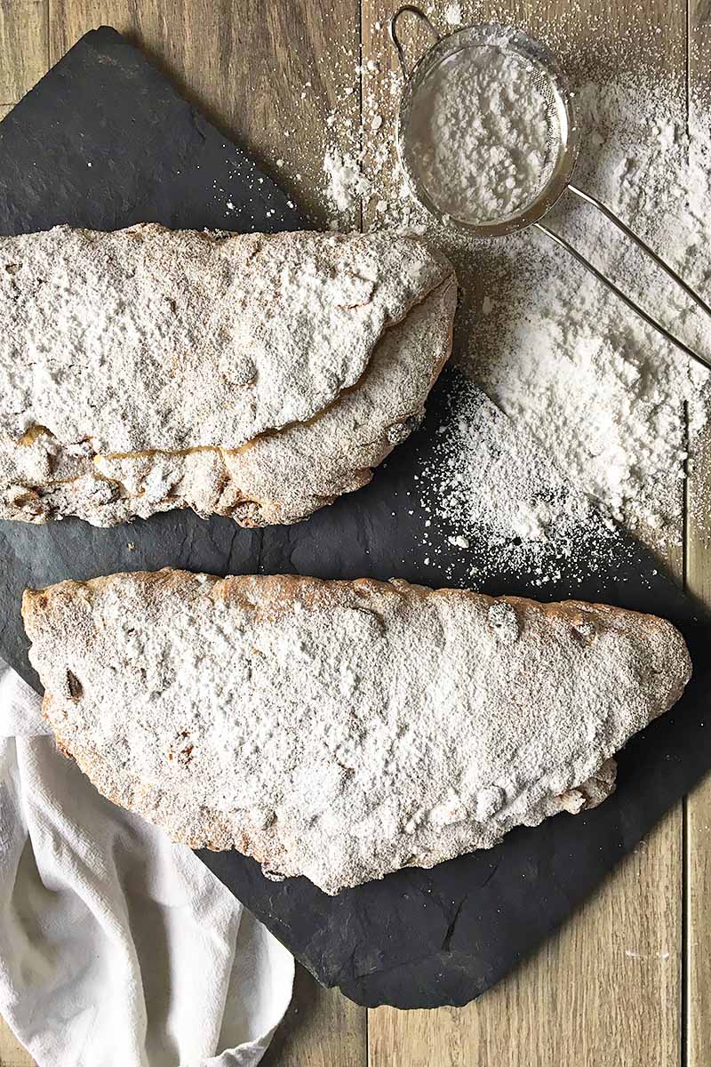 Vertical image of two whole loaves of stollen bread dusted in powdered sugar next to a small sifter with more powdered sugar.