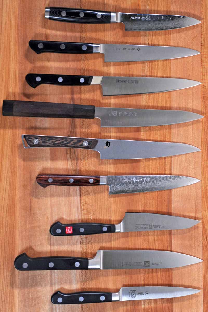 Top down view of all 9 of our reviewed petty and kitchen utility knives laid out on a maple butcher block surface.