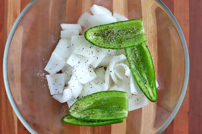 Overhead shot of chopped white onion and halved and seeded jalapenos topped with salt and pepper, in the bottom of a large glass mixing bowl, on a striped brown wood surface.