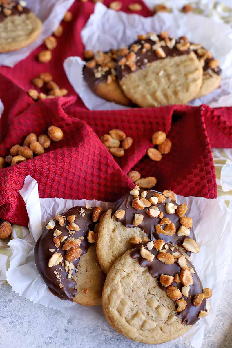 Chocolate peanut cookies arranged in groups of three on small pieces of parchment paper, on top of a gathered shiny red cloth with scattered honey roasted nuts.