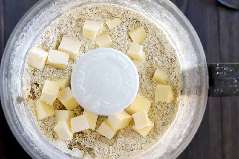 Overhead shot of a flour mixture topped with cubes of chilled butter in the canister of a food processor, on a dark brown wood background.