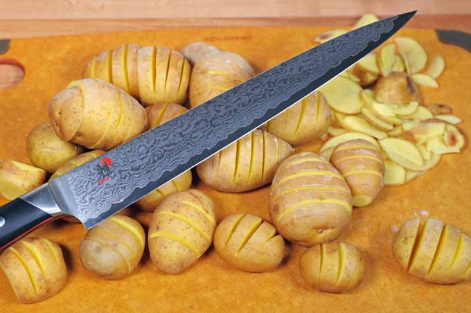 A Miyabi Japanese slicing knife is being used to cut slits along the length of small potatoes for a Hasselback-style recipe.