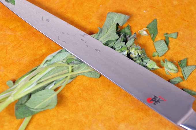 Sage is being chopped with a Japanese slicing knife on an Epicurean cutting board.