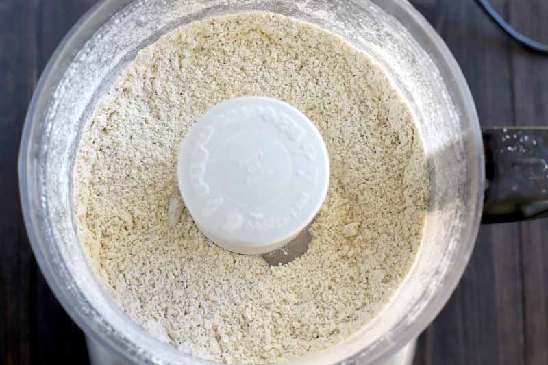 Overhead shot of a dry flour mixture in a white and clear plastic food processor, on a dark brown wood surface.
