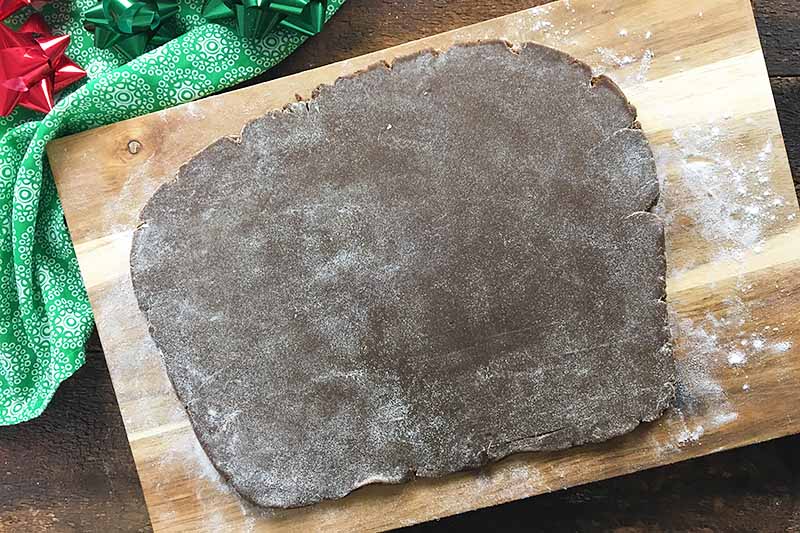 Horizontal image of rolled out dark brown dough on a wooden surface next to a green towel and Christmas decorations.