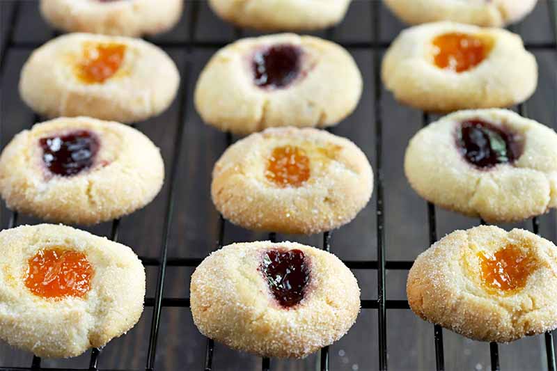 Jam-filled gluten-free almond baked goods filled with jam and coated with sugar are cooling on a metal wire rack, on a dark brown surface.