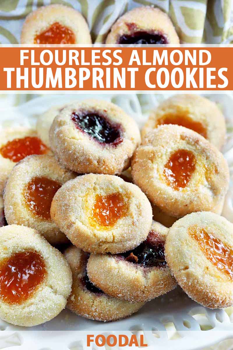 Vertical closeup of homemade almond thumbprint cookies filled with jam, arranged on a serving dish with a green and white cloth in the background, printed with orange and white text.
