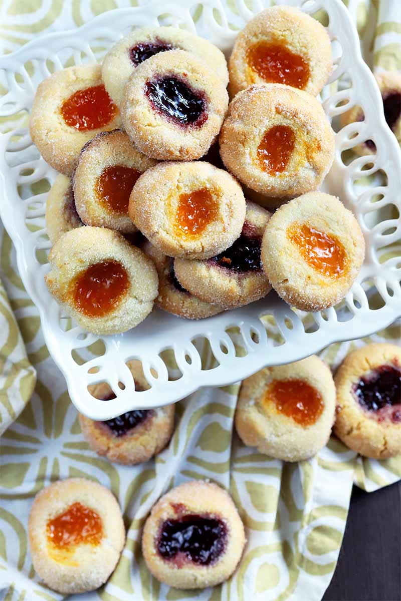 Overhead vertical shot of homemade flourless thumbprint cookies arranged on in a square white dish and scattered on a patterned cloth below, on a dark brown wood surface.