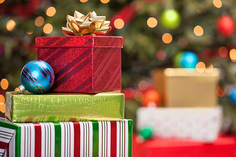 A stack of presents wrapped in holiday paper with a gold bow on top and a blue glass tree ornament, with more wrapped gifts and a decorated evergreen tree in soft focus in the background.