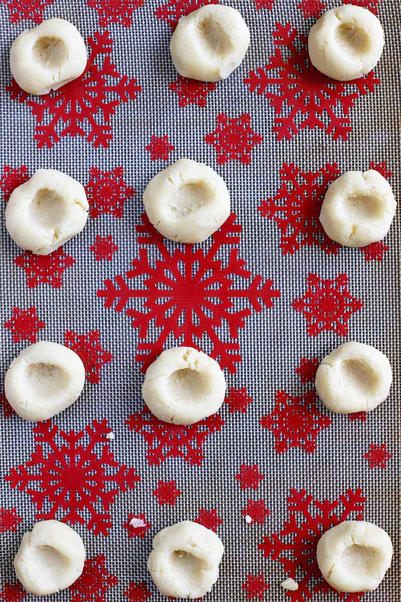 Top-down shot of cookie dough rolled into balls and pressed with a thumbprint indentation in the center of each, arranged in rows on a silicone pan liner with a red snowflake pattern.