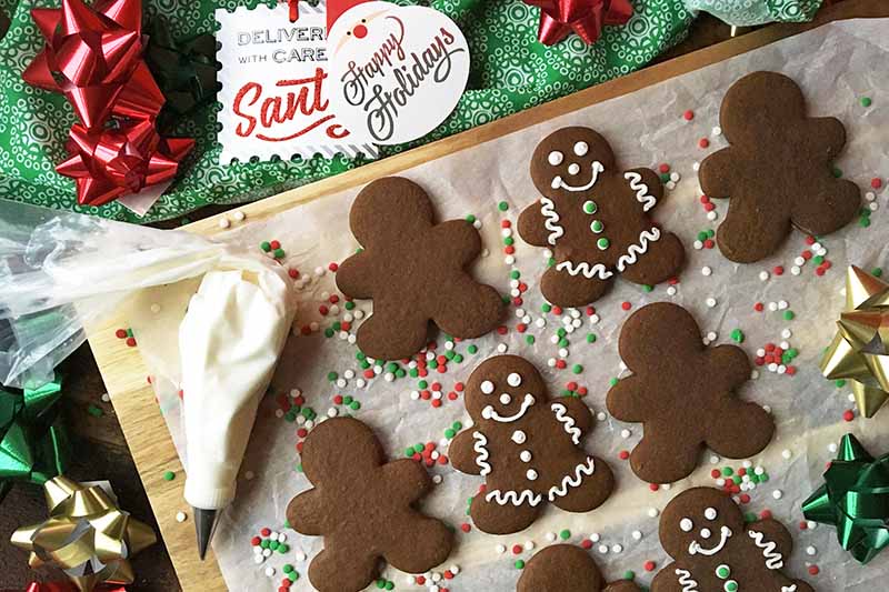 Horizontal image of decorated Christmas gingerbread cut-outs on a wooden board next to a piping bag and holiday decorations.