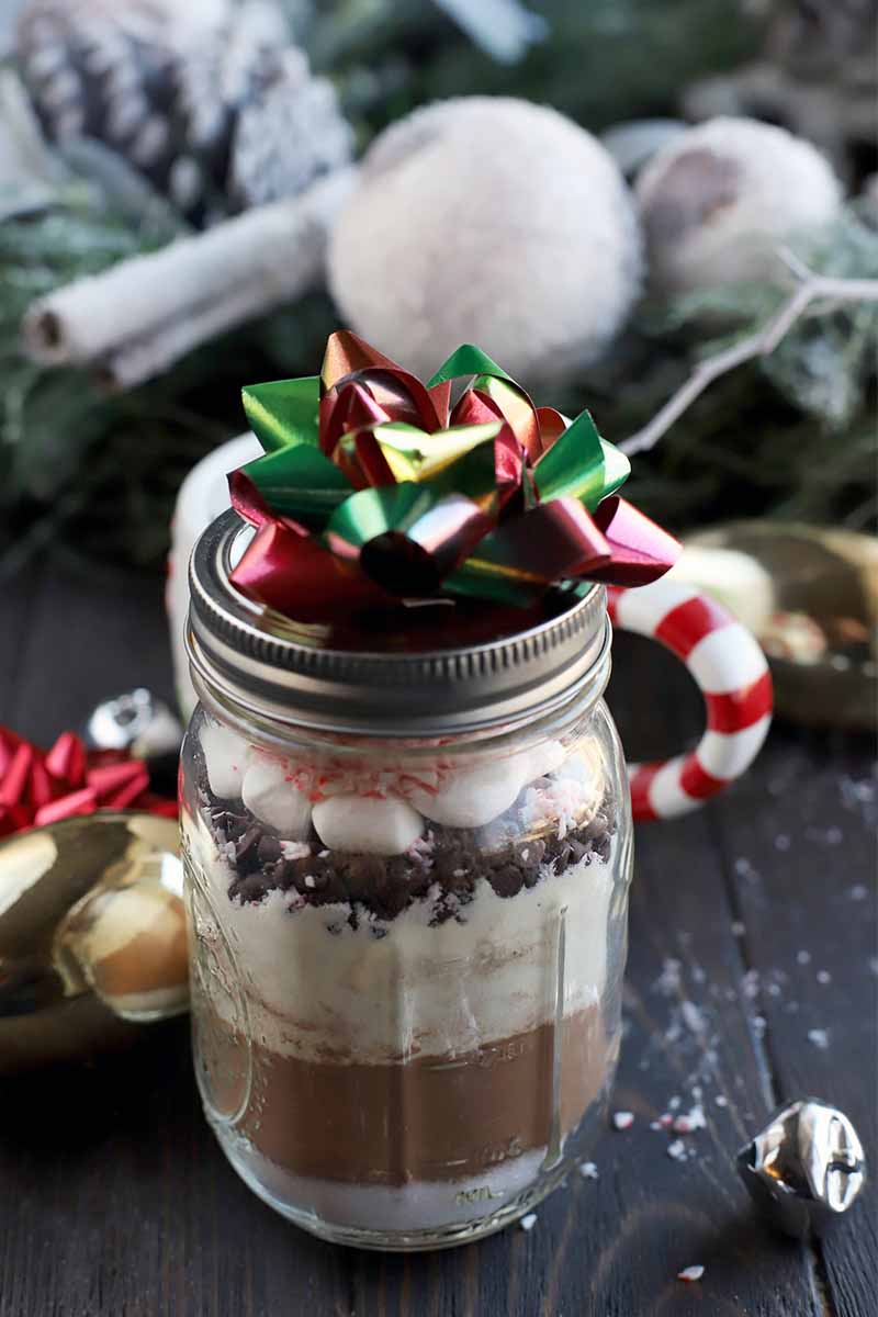 A jar of homemade hot chocolate mix topped with a bow is in the foreground, with holiday greenery and decorations in the background, on a dark brown wood table, with scattered candy cane crumbs.