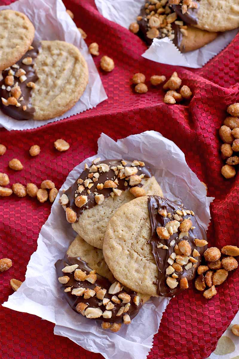 Chocolate-dipped peanut cookies are arranged in groups of three on small pieces of crumpled parchment paper on a gathered red cloth with scattered honey roasted nuts.