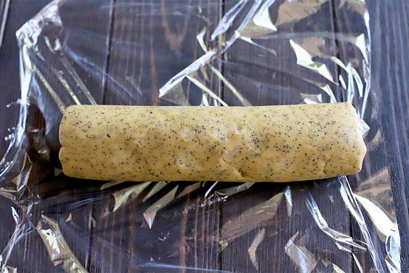 A log of cookie dough is resting on a shiny piece of plastic wrap, on a dark brown wood surface.