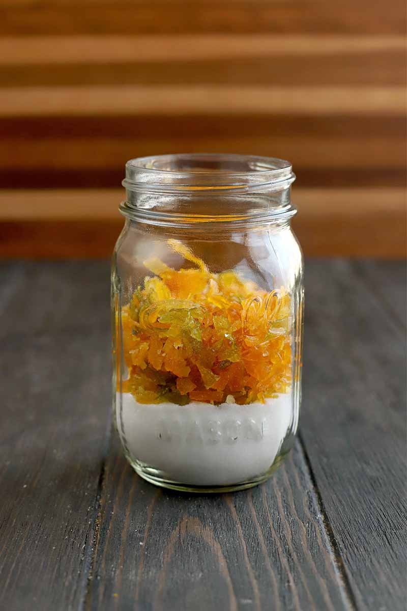 A mason jar filled with a layer of sugar at the bottom, topped with candied citrus peel, on a dark brown wood table against a brown striped wood backdrop.